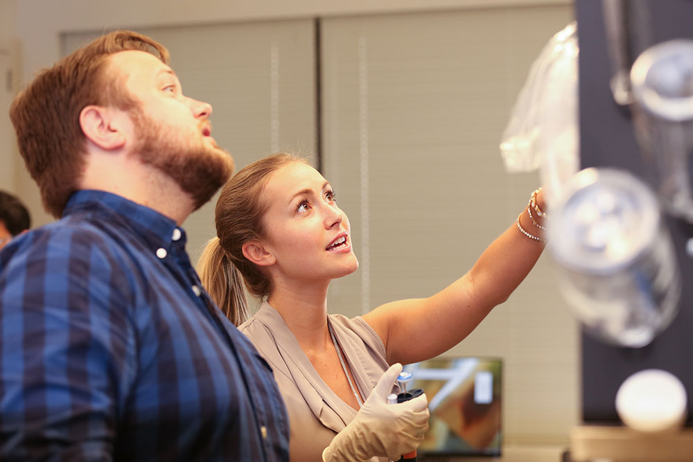 Michael Ludwig and Caitlin McMenamin, both students in the Anatomy PhD Program at Penn State College of Medicine, are seen at work in a lab in the Department of Neural & Behavioral Sciences in 2016. The two are looking downward and framed at right by clean glassware hanging on a lab wall.