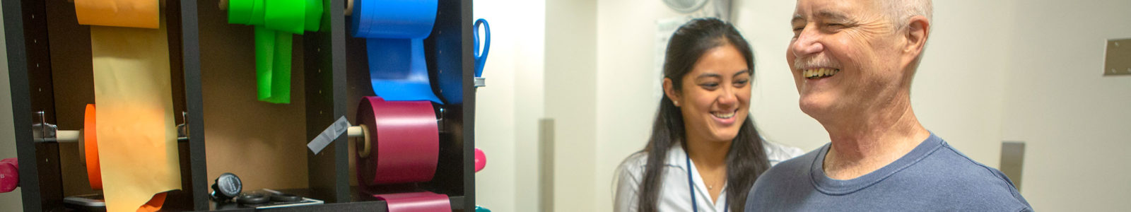 ONE Group staff member Melanie Potiaumpai, PhD, works with cancer patient Curt Chambers in the exercise room at Penn State Cancer Institute in 2018. Both are smiling and Chambers is pulling on a resistance band.