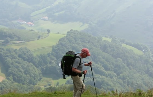 A photo shows a man with a backpack and two hiking poles, walking along a peak with a valley visible beside him. Trees and fields are visible below.