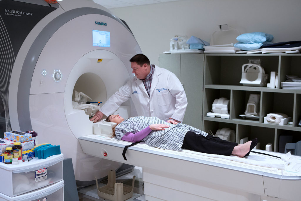 A doctor stands next to a person who is getting ready to slide into an MRI scanner. Both look relaxed.