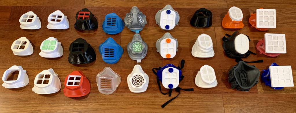 A collection of 27 colorful 3D printed protective face mask prototypes are arranged on a wood table during a meeting where clinicians were invited to test each model and provide direct feedback on fit, size and comfort.