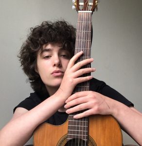 Gavi Kamen poses for a portrait holding a guitar upright in front of him