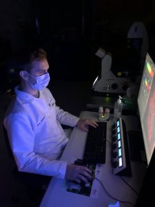 Dr. Greg Lambert, wearing a white coat, sits at a desk in front of a computer screen in the microscopy core facility.
