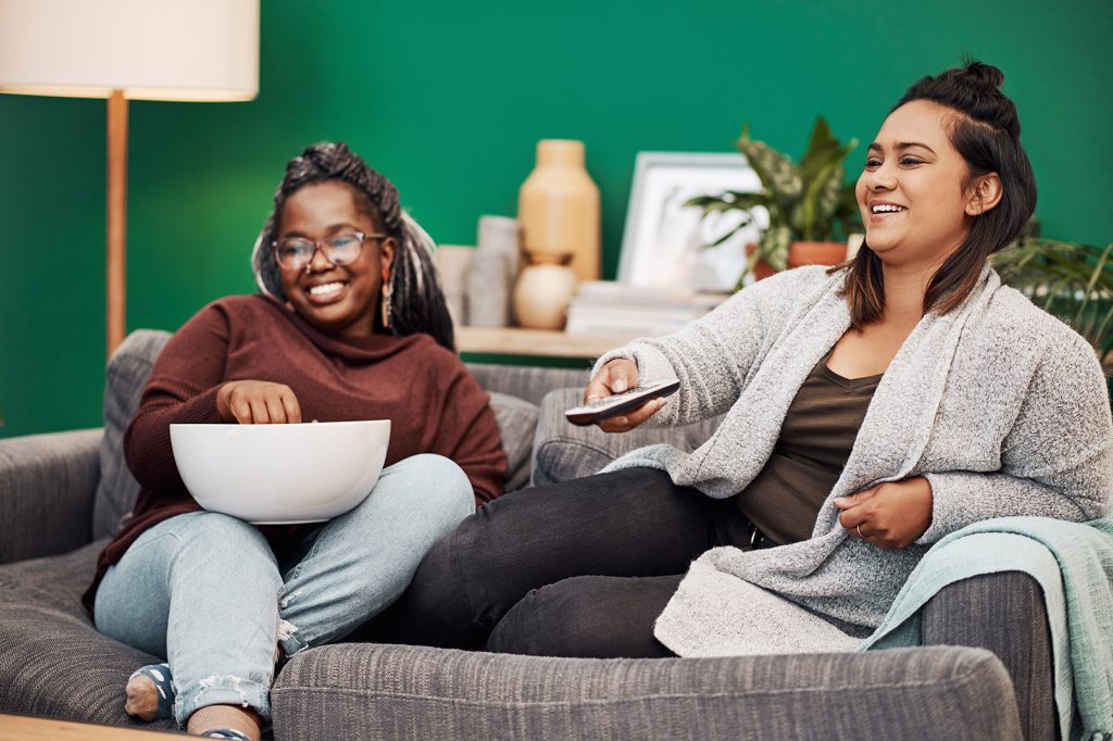 Two young women sitting on couch watching TV, out of frame, the woman on the left is holding popcorn and the woman on the right is using a remote control
