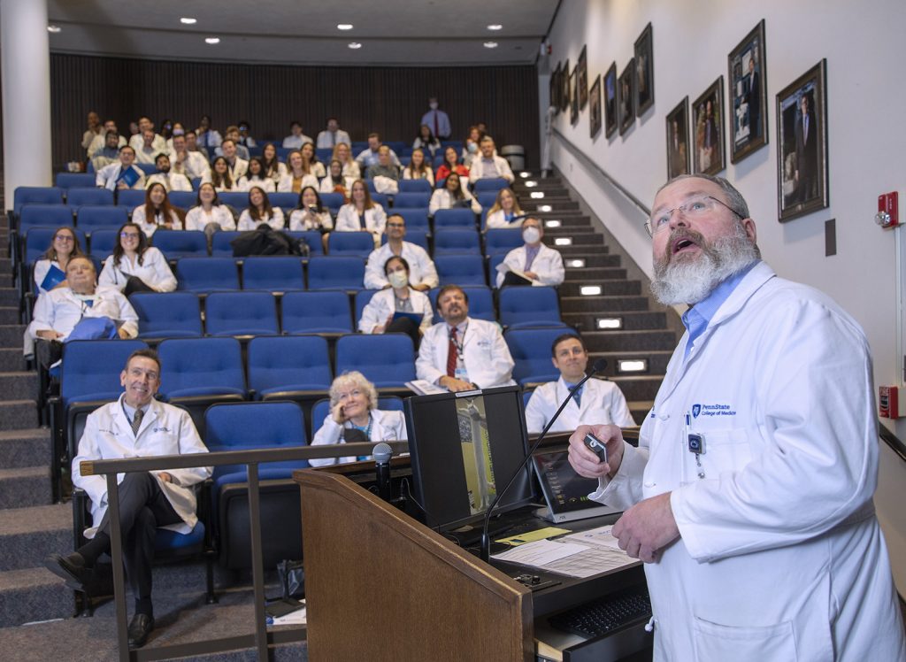 Dr. Ronald Miller in physician's white coat behind a lecturn looks back and above the camera while people in auditorium seats in the background laugh.