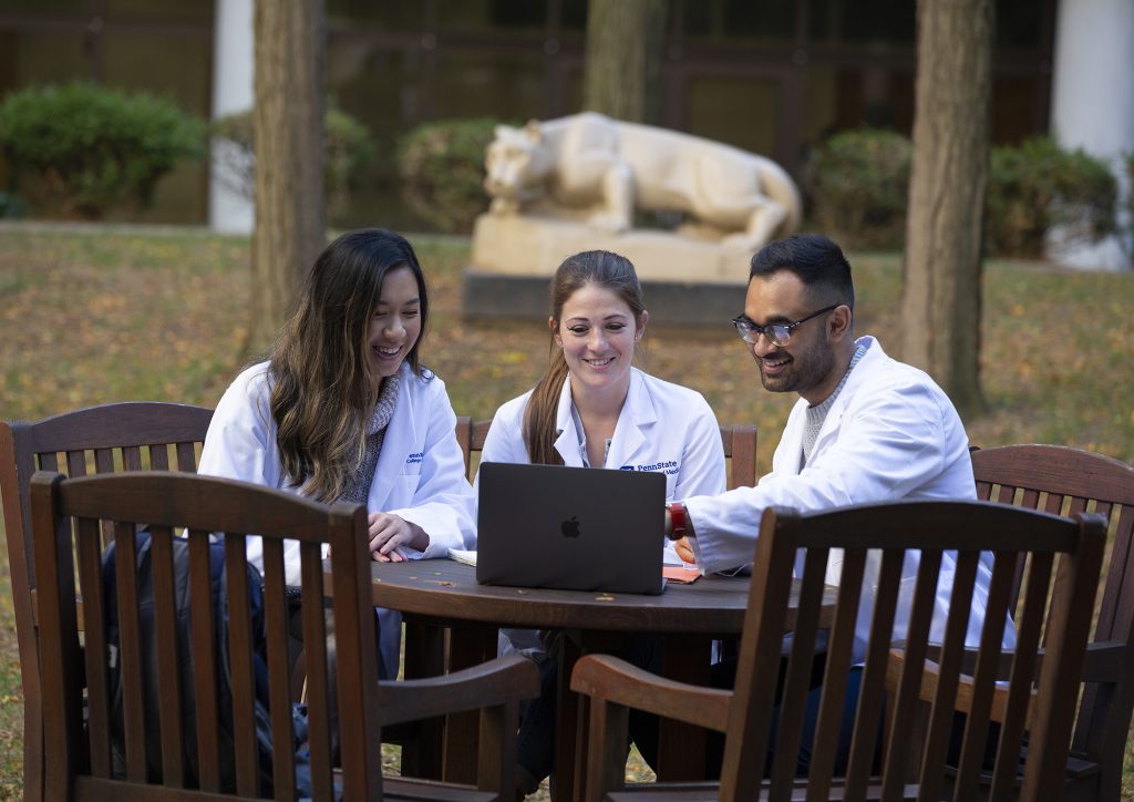 Three students at an outdoor courtyard table with a laptop in the middle of them and the Nittany Lion statue in the background.