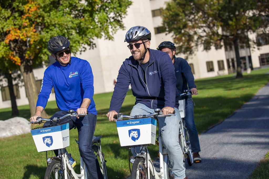 Three students, two in foreground and one behind, bicycling on bikes with a Penn State logo on the front and wearing bike helmets. 