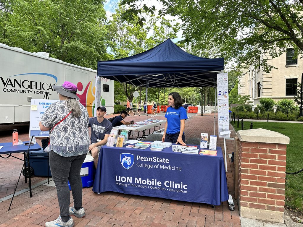 A woman in a Penn State College of Medicine shirt stands under a tent and behind a table with a LION Mobile Clinic Banner, smiling looking to the side
