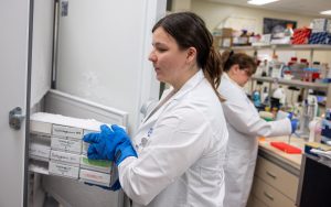 Erin King holding items in a lab, with a person working at a lab bench in the background