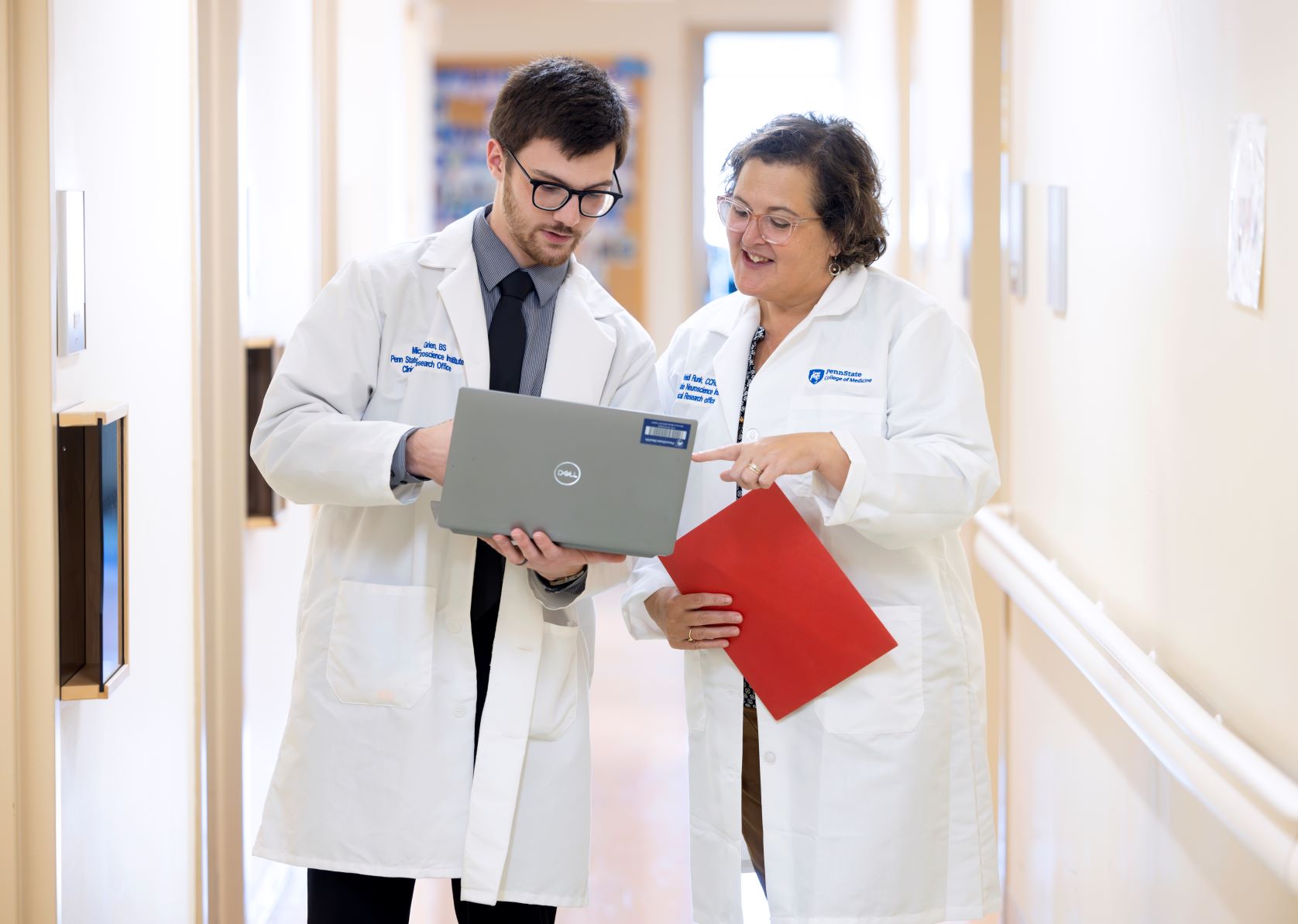 Micaiah Grien and Heidi Runk, both wearing white coats, stand in a hallway looking at a laptop Grien is holding.