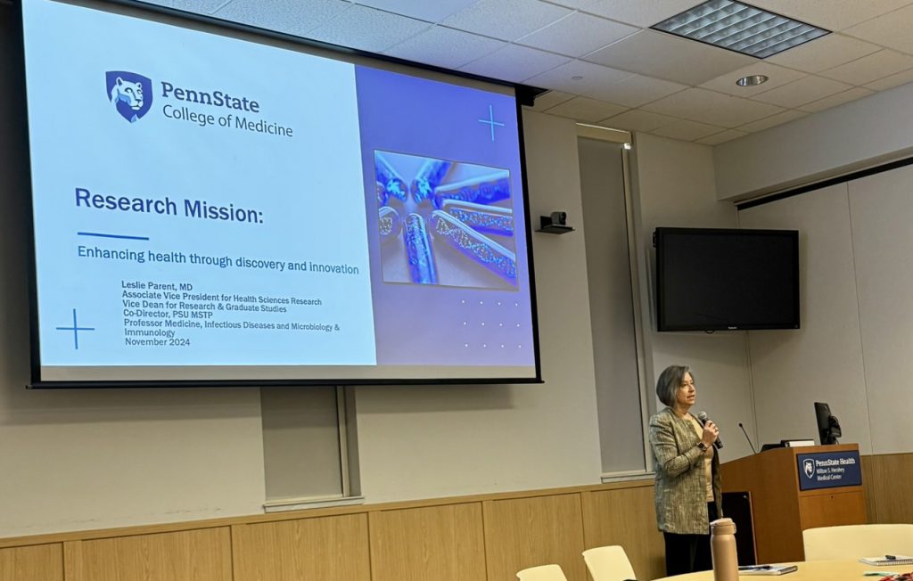 Dr. Leslie Parent holds a microphone and stands next to a lectern, with a large screen projecting a slide behind her and to her left.